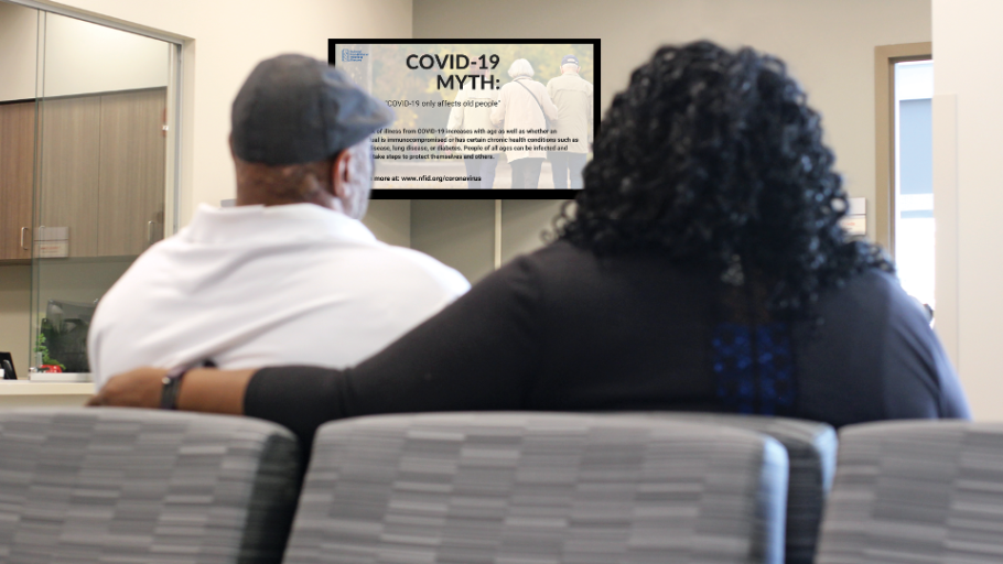 Two individuals sitting in grey chairs in a waiting room, viewing a tv screen that states "COVID-19"