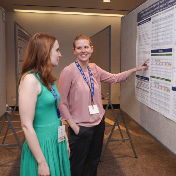 Two women stand in front of a poster, with one pointing.