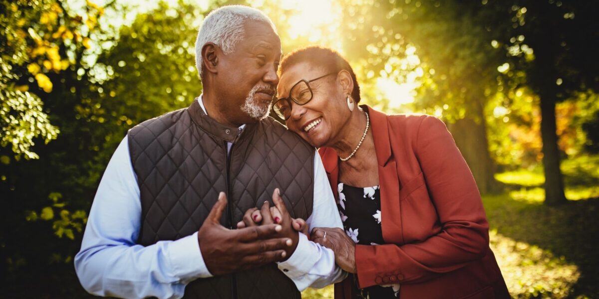 Una pareja negra mayor está de pie al aire libre en un parque iluminado por el sol, sonriendo y tomados de la mano