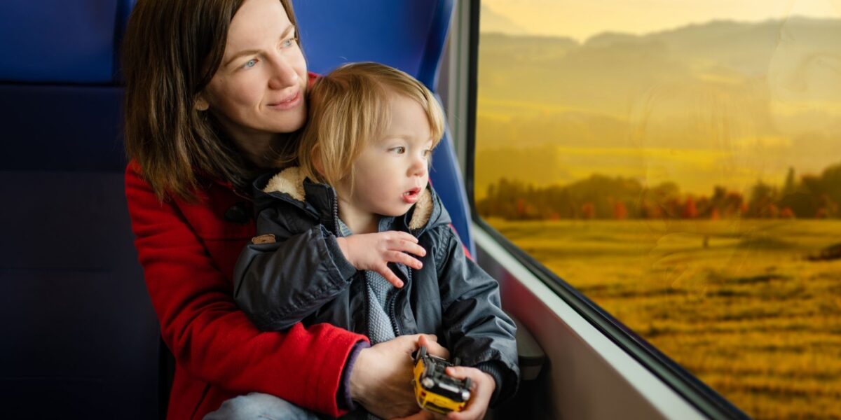 Una mujer y un niño pequeño se sientan juntos junto a la ventana de un tren, mirando un paisaje escénico del campo. El niño sostiene un coche de juguete mientras ambos miran la vista que pasa.