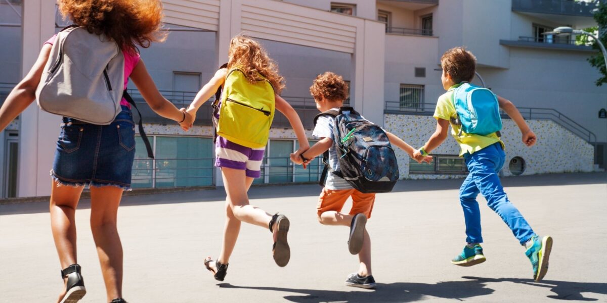 Four children wearing colorful backpacks run hand in hand across a courtyard toward a school building. The children are photographed from behind, with bright backpacks in gray, yellow, black, and blue
