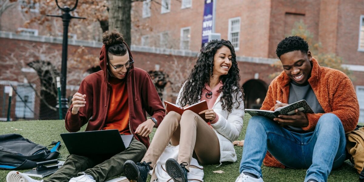 Tres estudiantes universitarios se sientan juntos en el césped del campus, sonriendo y estudiando con cuadernos y una computadora portátil frente a edificios académicos de ladrillo.