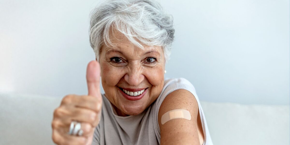 Smiling, silver-haired woman gives a thumbs up and shows the bandage from her vaccination