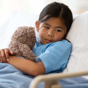 Young girl resting in a hospital bed while holding a stuffed animal, wearing a blue patient gown and covered with a blanket