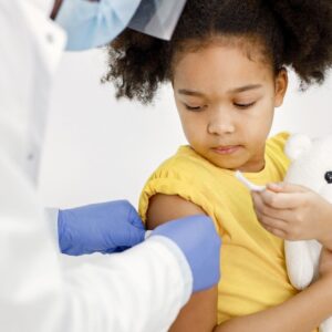 Young girl holding a stuffed animal while a healthcare professional wearing gloves puts a bandaid on her upper arm