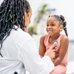 Smiling young girl sitting with a healthcare professional who listens to her chest with a stethoscope