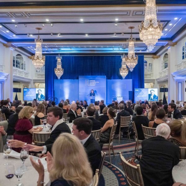 2024-nfid-awards-10 A woman stands at the podium of an elegant ballroom with crystal chandeliers and round tables of people eating dinner