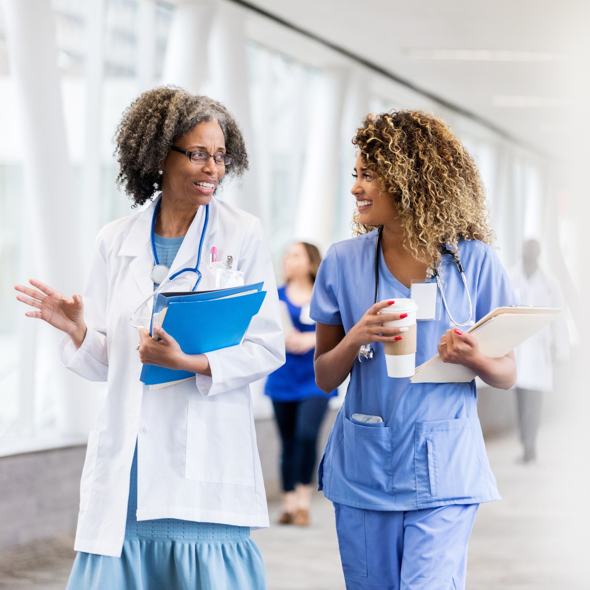 Two healthcare professionals walk side by side through a bright hospital corridor while smiling and talking. One wears a white lab coat and carries blue folders, while the other wears blue scrubs, carries a tablet and a coffee cup.