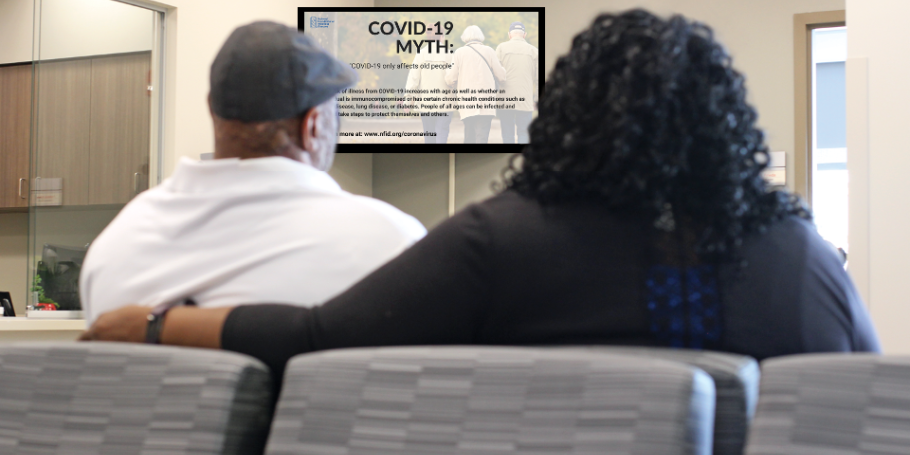 Two individuals sitting in grey chairs in a waiting room, viewing a tv screen that states "COVID-19"